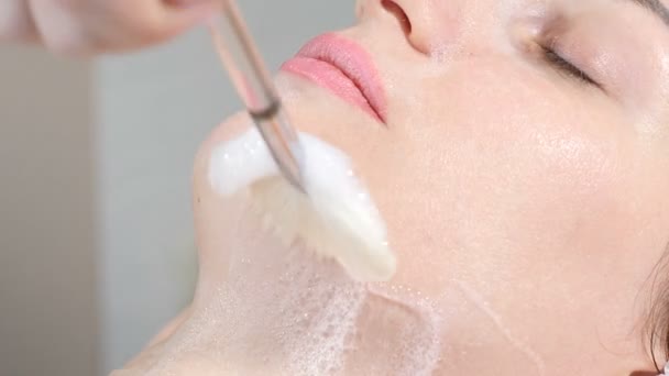 美容クリニックのコンセプトです。美容師のクローズ アップは、注射前に女性の顔にブラシで拡散洗浄マスクを手します。若い女性は、顔の手順を取得します。顔の若返り。4 k — ストック動画