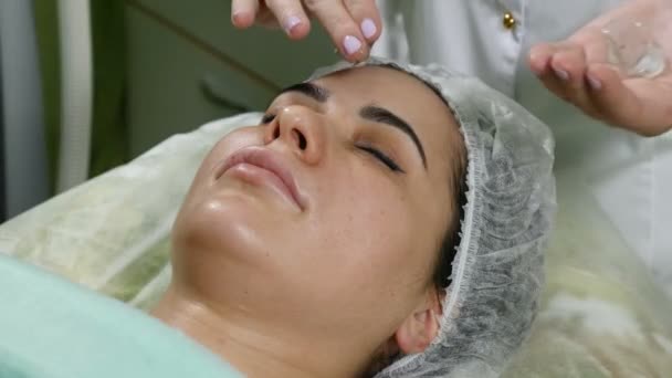 Концепция клиники красоты. Крупный план косметолога, намазывающего крем перед процедурой лица. Молодая женщина. Омоложение лица. 4k — стоковое видео