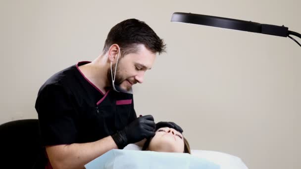 Salão de beleza. Esteticista masculino em luvas pretas fazendo procedimento de maquiagem permanente em sobrancelhas femininas. Gestos cosmetologistas masculinos sorridentes durante o procedimento — Vídeo de Stock