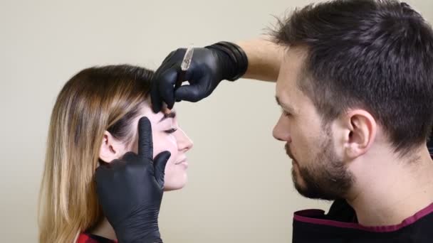 Salón de belleza. esteticista masculino en guantes negros aplicando delineador de cejas antes del procedimiento de maquillaje permanente. Mujer joven obtiene procedimiento de belleza facial. Rejuvenecimiento facial — Vídeos de Stock