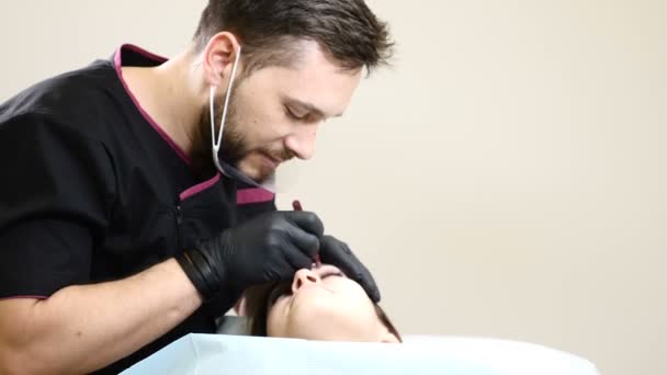 Schoonheidssalon. Mannelijke schoonheidsspecialiste in zwarte handschoenen maken van permanente make-up procedure op vrouwelijke wenkbrauwen. Lachende mannelijke schoonheidsspecialist gebaren tijdens procedure — Stockvideo