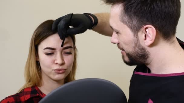 Beauty salon. Final touch.Male beautician in black gloves making permanent makeup procedure on female eyebrows. Female client holds a mirror — Stock Video