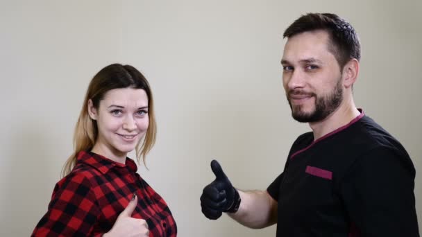 Portrait of happy female client and male beautician in healthcare clinic cabinet of beauty salon. Male cosmetologist in black gloves making permanent makeup procedure on female eyebrows. Thums up — Stock Video