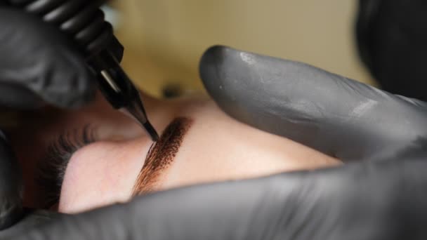 Klinika urody. Zbliżenie mężczyzna kosmetyczka w czarne rękawiczki Dokonywanie procedury Makijaż permanentny na kobiece brwi. Młoda kobieta dostaje procedury piękno twarzy. Odmłodzenie twarzy. Przy użyciu maszyny do tatuażu — Wideo stockowe
