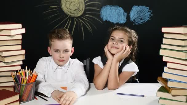 回到学校的概念。男孩和女孩坐在一张桌子上, 里面堆着书和黑板, 后面画着图画。孩子们玩得开心。女孩拥抱她的同学。4k — 图库视频影像