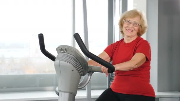 Retrato de una anciana activa en el gimnasio haciendo entrenamiento cardiovascular en una bicicleta estática. Vieja sana y activa. Personas mayores activas. Una anciana en la sala de fitness. Disparo en 4k — Vídeo de stock