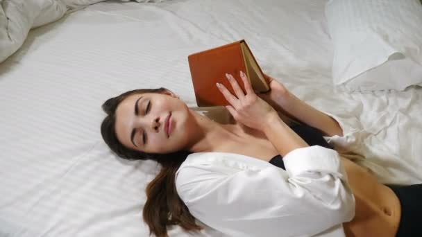 아름다운 아가씨가 큰 침대에 누워 있는 동안 손에 쓰는 쪽지를 들고 집에서 쉬고 있다. 하얀 침대에 누워 눈을 감고 누워 있는 호텔 방의 매력적 인 여성. 출장이요. 4k — 비디오