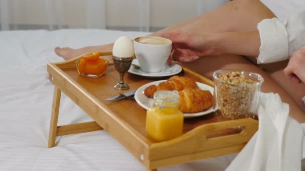 Closeup shot of young woman drinking coffee in cosy hotel room in the morning. Table tray on white bedding with egg, cup, croissants. jam, honey, muesli and carrot juice. Healthy breakfast. Shot in 4k — Stock Video