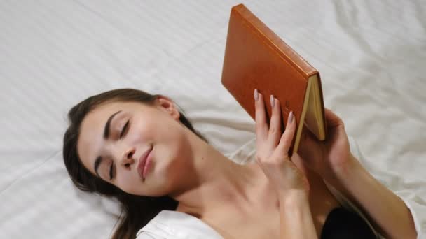 Egy fiatal, csinos nő fekszik az ágyában egy zárt füzetben a kezében. Pihentető, vonzó hölgy fehér ágyon reggel. 4k — Stock videók