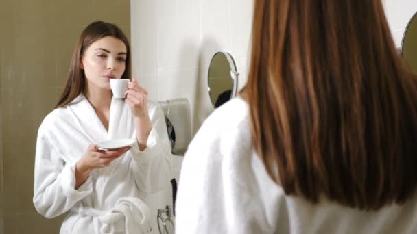 Молодая привлекательная женщина в белом халате пьет кофе перед зеркалом в ванной. Запись с заднего вида. Женщина смотрит на себя в зеркало ванной комнаты и улыбается, наслаждаясь утром — стоковое видео