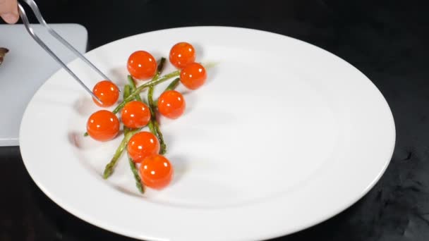 Cocina de chef en la cocina del restaurante. Poner cereza de tomate y espárragos con pinzas en plato blanco. Sirviendo platos. Buena cocina. En cámara lenta. Hd — Vídeos de Stock