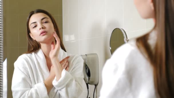 Porträt einer jungen Hotelbesucherin im weißen Bademantel, die sich selbst im Spiegel betrachtet. Süße Frau in ihren Zwanzigern, die ihr Gesicht und ihre Haare mag, selbstbewusst und zufrieden mit ihrem Aussehen. Schuss in 4k — Stockvideo