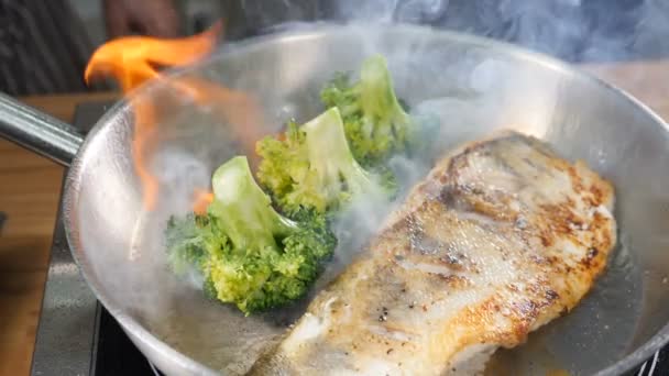식당 주방에 있는 프라이팬에 야채를 곁들여 생선을 요리한다. 닫아. 느린 동작으로 발사되는 가스 버너입니다. 불꽃 속의 브로콜리. 완벽 한 음식 영상이야. HD 주사 사진 — 비디오