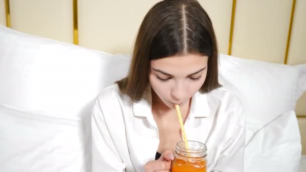 Портрет привлекательной молодой женщины, улыбающейся в камеру и пьющей морковный сок через трубочку. Девочка на кровати с белыми подушками на фоне. Завтрак в постель утром. Выстрел в 4k — стоковое видео