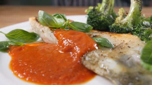 Здорова страва з морепродуктів, приготована шеф-кухарем у ресторані "Bourmod". Делікатний соус наливають на білу тарілку зі смаженим рибним філе та брокколі. Повільний рух. Постріл в HD — стокове відео