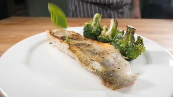 Touche finale en cuisine. Le plat du restaurant est décoré et servi avec des feuilles vertes de potherbs. Ferme là. Feuilles tombant sur une assiette blanche avec du poisson et des légumes frits. Hd — Video