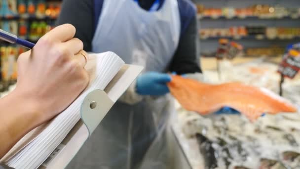 Qualitätskontrolle der Lebensmittelversorgung. Nahaufnahme von weiblichen Händen, die Notizen schreiben, während sie Produkte für Restaurants im Supermarkt auswählen. Männliche Hände zeigen Lachsfilet. Schuss in 4k — Stockvideo