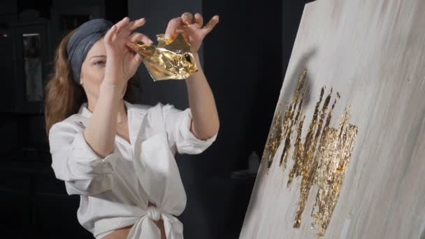 Attraktiv kvinnlig konstnär som applicerar guldblad på sitt konstverk. Konstnären dekorerar bilden med små ark av guld. Konstskola, kreativitet och människors koncept. Sakta i backarna. Skjuten i hd — Stockvideo
