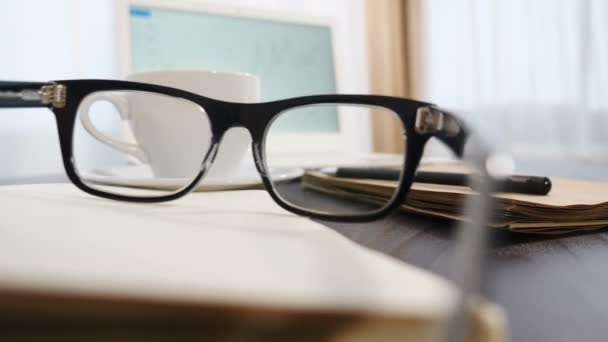 Concepto de negocio. De cerca. Las gafas se ponen en el bloc de notas. Cuaderno blanco y una taza de café de fondo. Gafas, bolígrafo y cuaderno vacío abierto en el escritorio de la oficina. Disparo en 4k — Vídeos de Stock