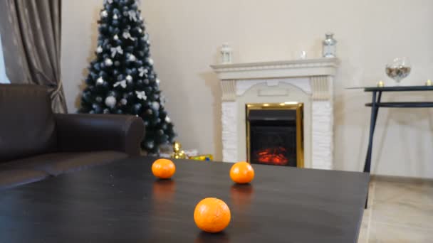 Preparação de Ano Novo e Natal. As mãos femininas colocam um vaso de vidro cheio de tangerinas maduras na mesa preta. Árvore de Ano Novo perto do lugar de fogo e decorações no fundo. Tiros em 4K — Vídeo de Stock