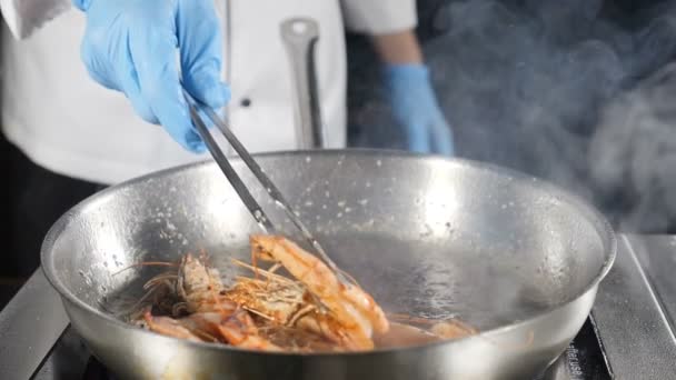 Koch braten Tigergarnelen und bewegen eine der Garnelen mit einer Kulinarikzange. Restaurantessen, das von Meisterkoch in blauen Handschuhen zubereitet wird. Zeitlupe. Nahaufnahme. hd video — Stockvideo
