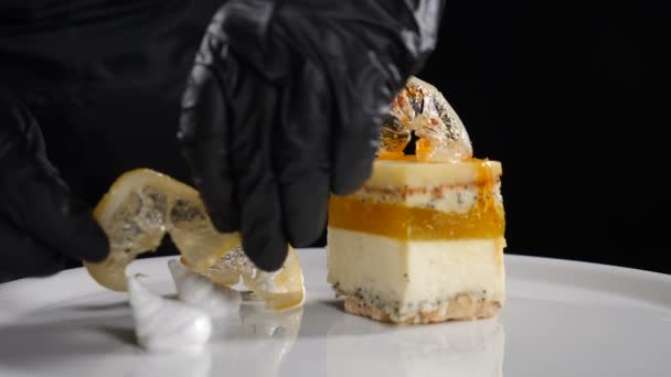 Preparation of delicious dessert at luxury restaurant. Chef in black gloves putting dried sliced sugar lemon on white plate with tasty pastry with layers. Close-up. Slow motion. Full hd — Stock Video