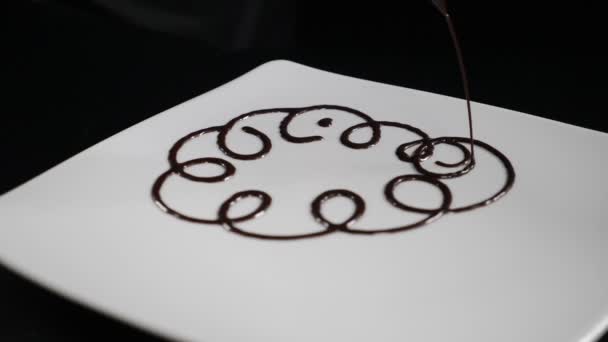 Processo de cozinhar e servir sobremesa no restaurante. chocolate derretido é derramado sobre placa branca em círculos em câmera lenta. Foto de perto de como um chef de pastelaria prepara uma sobremesa na cozinha do restaurante — Vídeo de Stock