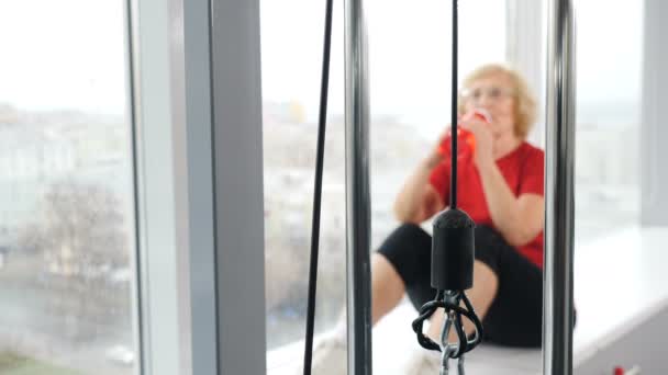 Senior active caucasian woman sitting on window sill and drinking water from bottle after training in gym, Retired woman enjoying old years. 4k footage — Stock Video