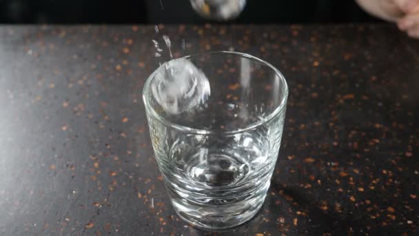 Top view. Close-up shot of ice cubes falling into empty clear glass for whisky or brandy or bourbon on dark bar counter, slow motion. Empty glass with ice cubes on wooden table on a black background — 비디오