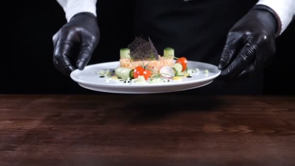 Comida saludable y buena cocina de restaurante. Chef en guantes negros sosteniendo plato con sarro cocido sobre fondo negro. Cook presenta su plato. Full hd — Vídeos de Stock