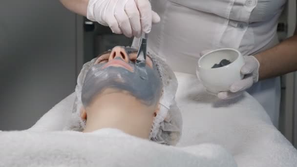 Jovem mulher bonita recebendo procedimentos no salão de beleza. Jovem mulher bonita deitada no sofá. Esteticista aplicando máscara creme facial no rosto. 4k vídeo — Vídeo de Stock