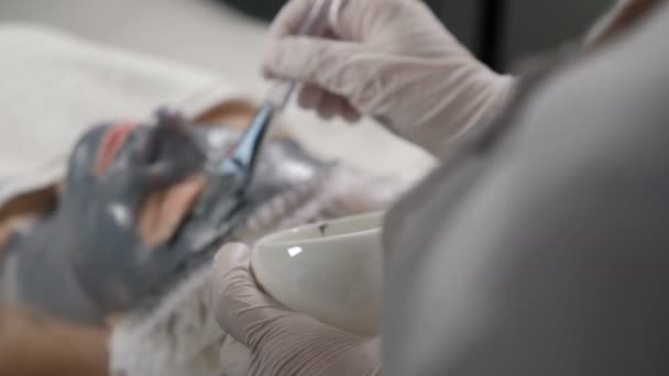 Cosmético aplicando mascarilla facial. Vista desde atrás. Primer plano de la joven atractiva en el salón de belleza. Procedimiento cosmético. Disparo en 4k — Vídeos de Stock