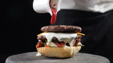 Chef preparing juicy burger and pours spicy sauce on it making burgers at fast food restaurant, Slow motion. Chef assembling a burger on black background. Fast food, unhealthy food concept. Full hd