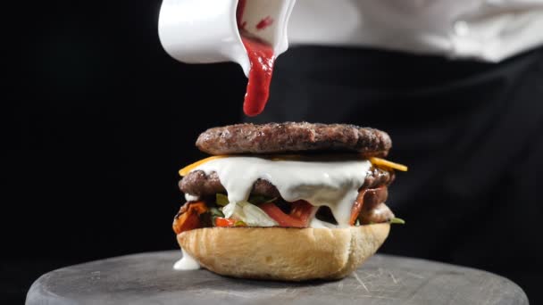 Chef menyiapkan burger juicy dan menuangkan saus pedas di atasnya membuat burger di restoran cepat saji, Lambat gerak. Koki merakit burger dengan latar belakang hitam. Makanan cepat saji, konsep makanan tidak sehat. Full hd — Stok Video
