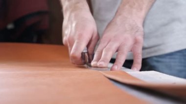 Hands of skilled leather craftsman using plastic pattern and leather knife to cut out part from piece of red leather, close-up shot. Industrial knife man cuts the work piece from the skin. 4 k footage