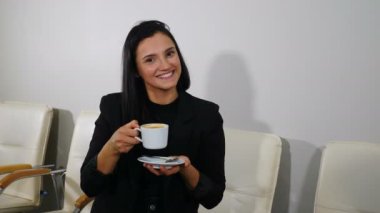 Medium shot of happy young smiling woman enjoying gorgeous coffee at office waiting for appointment. Girl waiting for health care procedure in clinic lobby. Shot in 4k