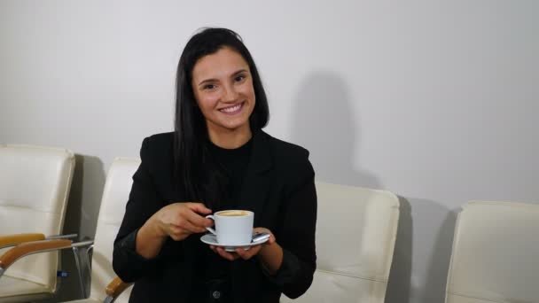 Happy girl drinking coffee and relaxing in office smiling and looking at camera. smiling woman enjoying gorgeous coffee at office waiting for appointment. Girl waiting for health care procedure in — Stok video