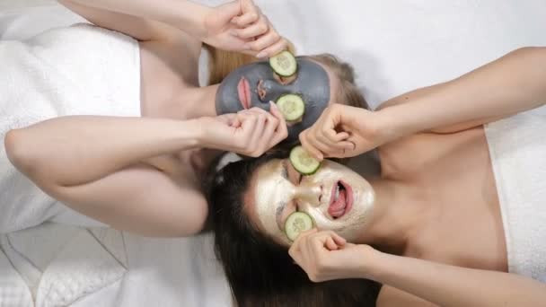 Des amies souriantes s'amusent avec du concombre tranché tout en appliquant un masque en argile, prises isolées sur fond blanc au salon de beauté. Temps de spa à la douche nuptiale. Vue de dessus. 4k — Video