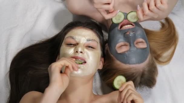 Sonrientes amigas que se divierten con pepino en rodajas mientras aplican mascarilla de barro, tiro aislado sobre fondo blanco en el salón de belleza. Hora del spa en la ducha nupcial. Vista superior. 4k — Vídeo de stock