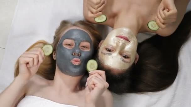 Top view on pretty ladies with dark and bright beauty cosmetology masks on faces on white floor and sincerely laughing holding sliced cucumbers. Wellness, spa, skincare and bachelorette party or — Stock Video