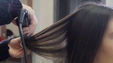 Hairstylist combing and straightening hair with hair iron in hairdressing salon. Close-up. Hairdresser doing female hairstyle with hot tongs in barbershop. Beauty and style concept. 4 k footage