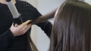 Process of curling long hair with hair tongs in modern beauty studio. Close-up shot of female hairstylist curling hair using hair iron. Hairstylist doing stylish hairdo. Macro. 4k footage