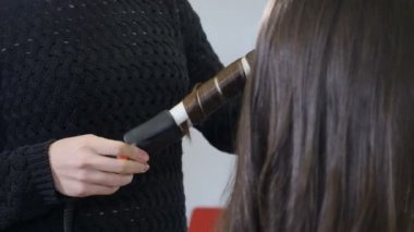 Beauty salon concept. Young woman while straightening hair. Hairstylist using hair iron and comb for woman hairstyling in hairdressing studio. Hairdresser creating female hairdo with tongs. 4k video