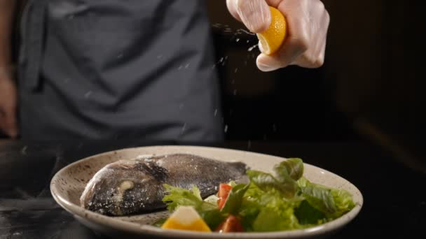 Zeitlupe zeigt, wie der Koch eine halbe geschnittene Zitrone über den Fisch drückt. Fisch mit grünen Blättern und Zitronenstück auf Teller im Restaurant serviert. Mann übergießt Fisch mit Zitronensaft und drückt Scheibe — Stockvideo