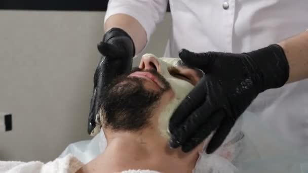 Concept of beauty salon and spa for men. Beautician applying facial cosmetic mask massaging with gloved hands. Spa therapy for handsome man getting rejuvenating facial treatment. 4 k footage — Αρχείο Βίντεο