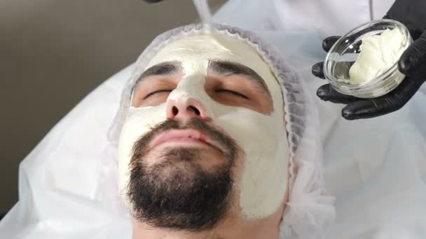 En la clínica de belleza. Cosmetólogo está aplicando mascarilla hidratante blanca en la piel de la cara masculina. Terapia de spa para hombre guapo recibiendo mascarilla facial. 4 k vídeo — Vídeos de Stock