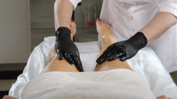 Primer plano de la esteticista femenina en guantes negros haciendo masaje y aceite de lavado corporal para una mujer joven. Preparación del cuerpo para envolverlo en Spa. Masaje en el salón de spa. 4 k vídeo — Vídeos de Stock