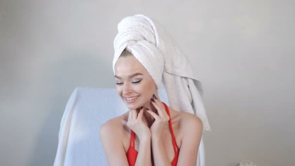 Vídeo retrato de uma jovem mulher de roupão de banho, com toalha branca na cabeça fechando os olhos com as mãos e olhando para a câmera. posando. Menina bonita sentada na cadeira no centro de spa de luxo. 4 k vídeo — Vídeo de Stock