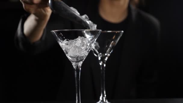 Кубики льоду потрапляють у порожнє скло при повільному русі. Близько. Жінка-бармен робить коктейлі на темному тлі. Концепція нічного клубу та бару. Повний HD — стокове відео