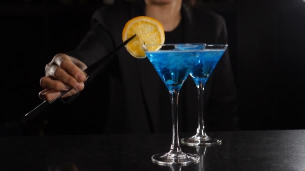Kant-en-klaar cocktail is op de bar in de nachtclub. Vrouwelijke barman versiert cocktail met blauwe vloeistof met schijfje citroen. Barmeid Decoratie Alcoholische Cocktail Vermouth Martini met verse citroen. Langzaam. — Stockvideo